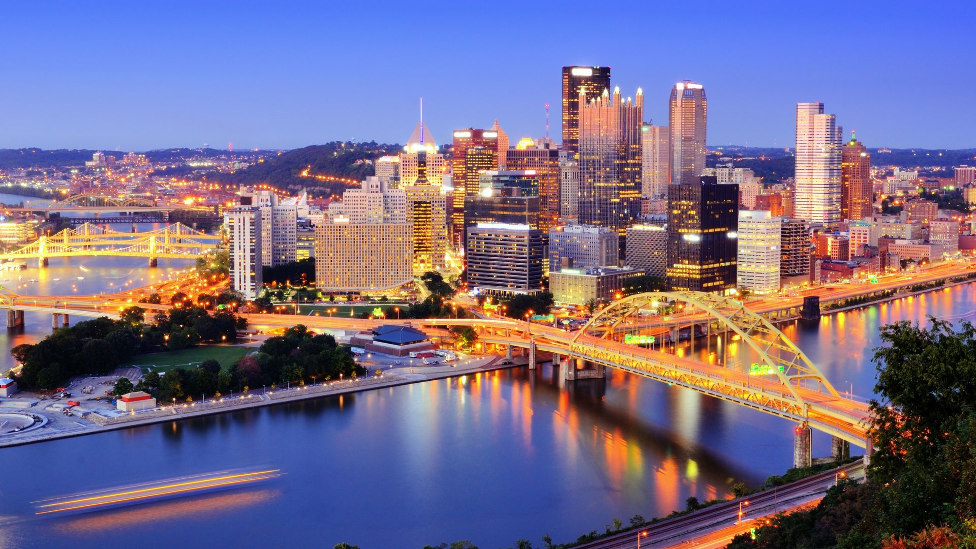 Night view of Pittsburgh skyline, lit buildings and bridges over a river, with lights reflecting in the water.
