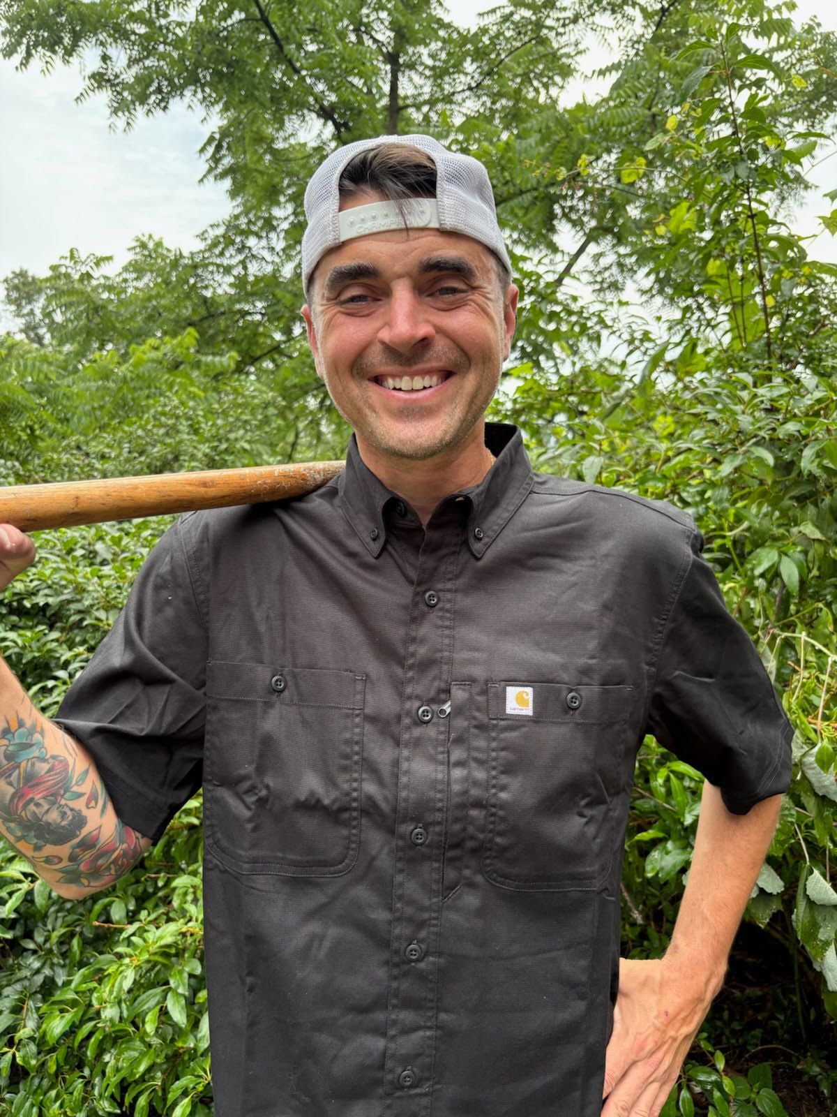 Man in black shirt and cap smiling, holding wooden pole, outdoors with greenery.