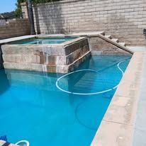 Blue swimming pool with a raised spa and a floating hose, against a brick wall.