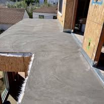 Fresh gray concrete rooftop walkway beside unfinished wooden structures and a view of nearby houses.