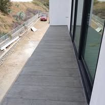 Narrow gray deck beside a building on a hillside, with a parked red vehicle below.