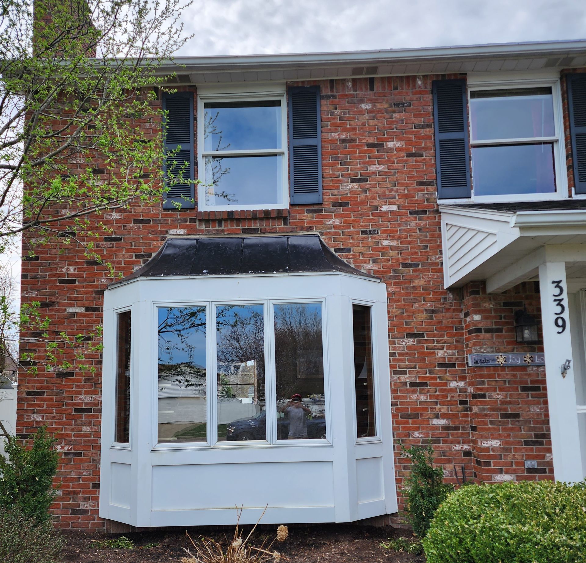 Two-story red brick house exterior with a white bay window on the ground floor and white shutters on the upper windows.