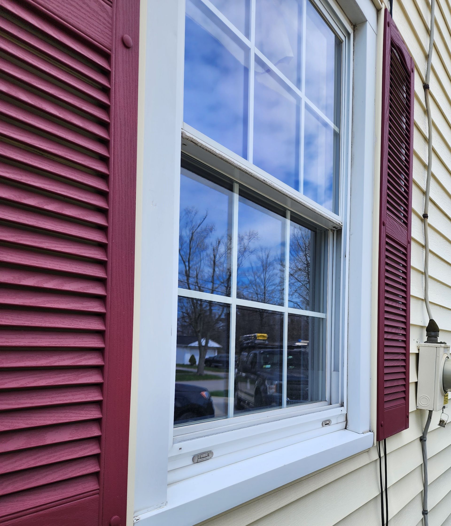 A double-hung window with a white frame and maroon shutters set into a cream-colored, vinyl-sided house exterior.