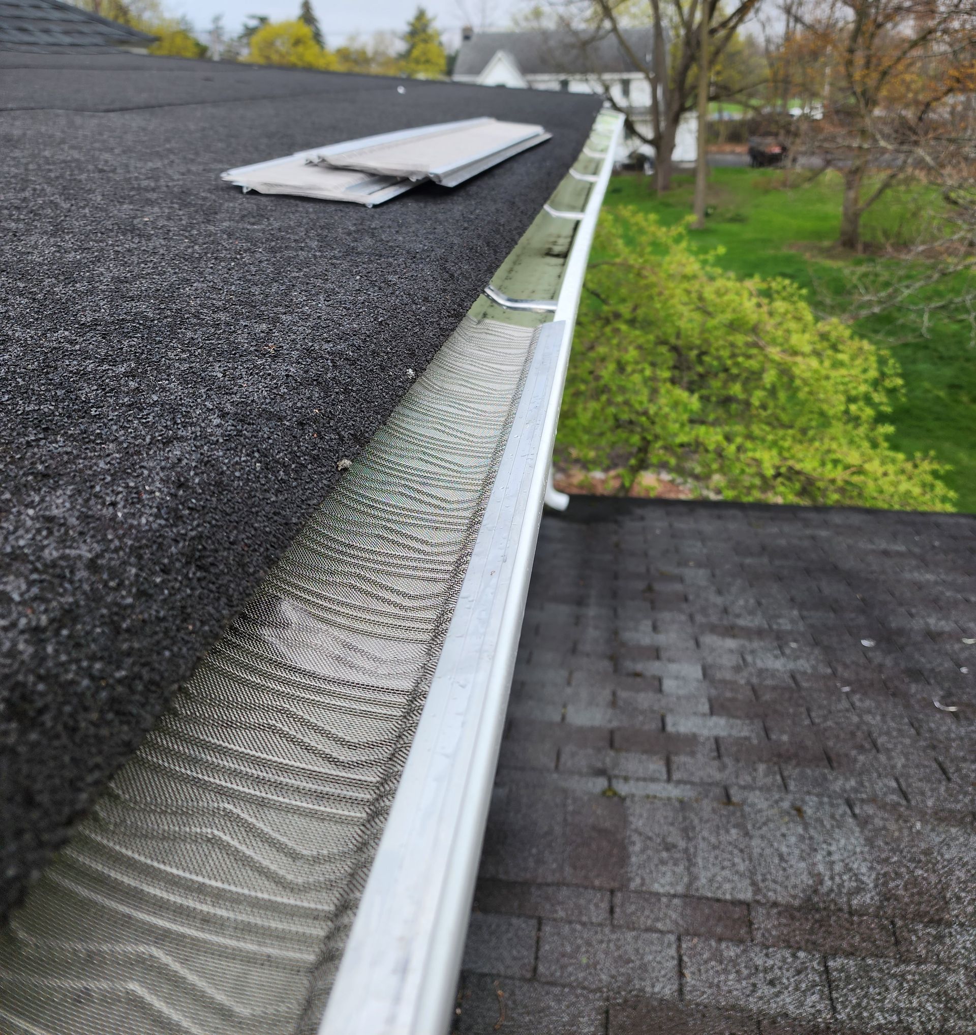 A white metal gutter with a mesh guard runs along the edge of a dark shingled roof, with an extra piece of guard on top.