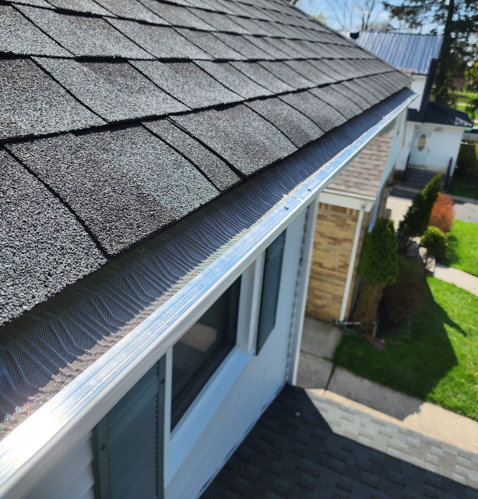 A high-angle view of a house roof with grey shingles and a silver gutter guard installed along the edge.
