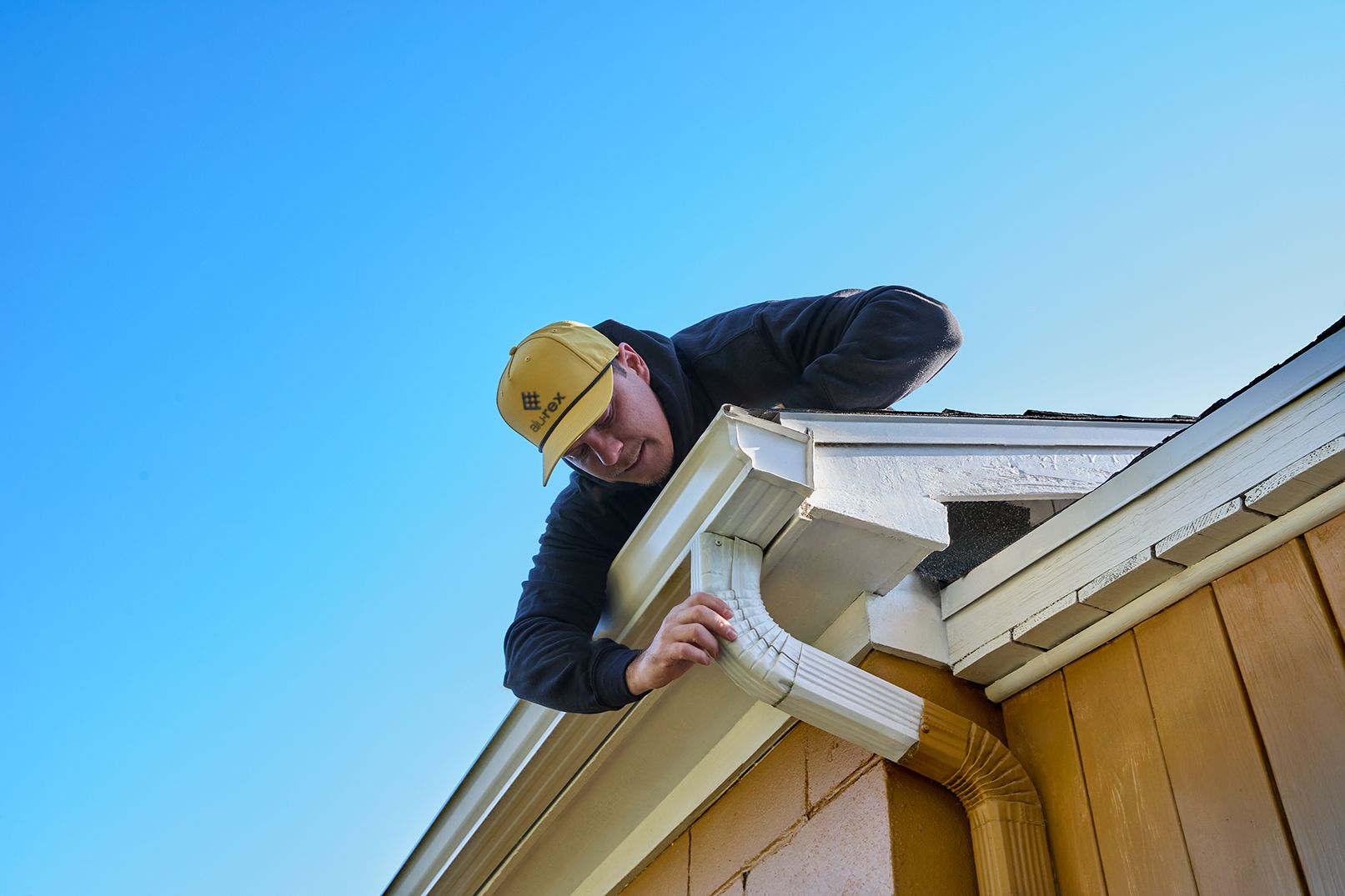 A worker on a ladder installs or repairs a gutter on the side of a tan house with a snow-covered roof.