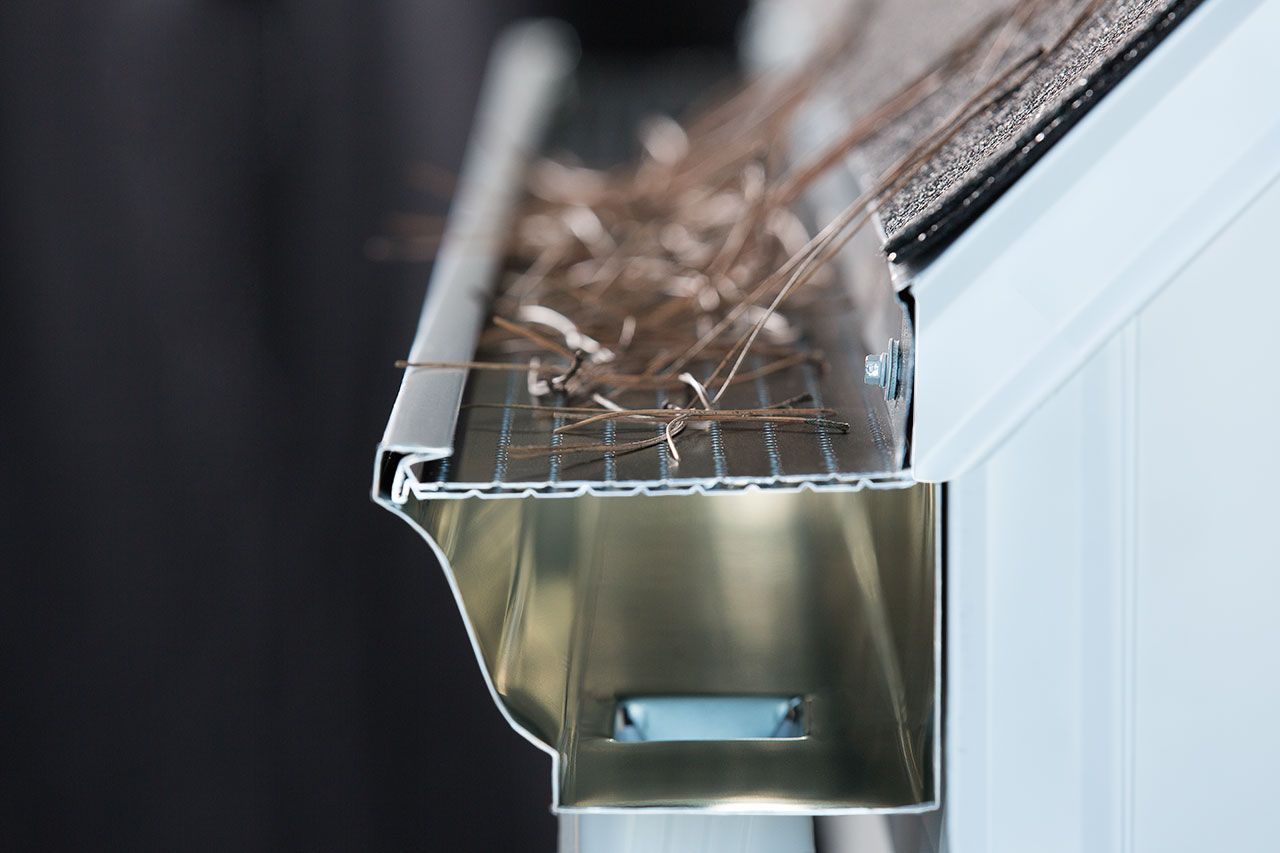 A close-up of a brown roof gutter and downspout installed under a tiled roof overhang against a clear blue sky.