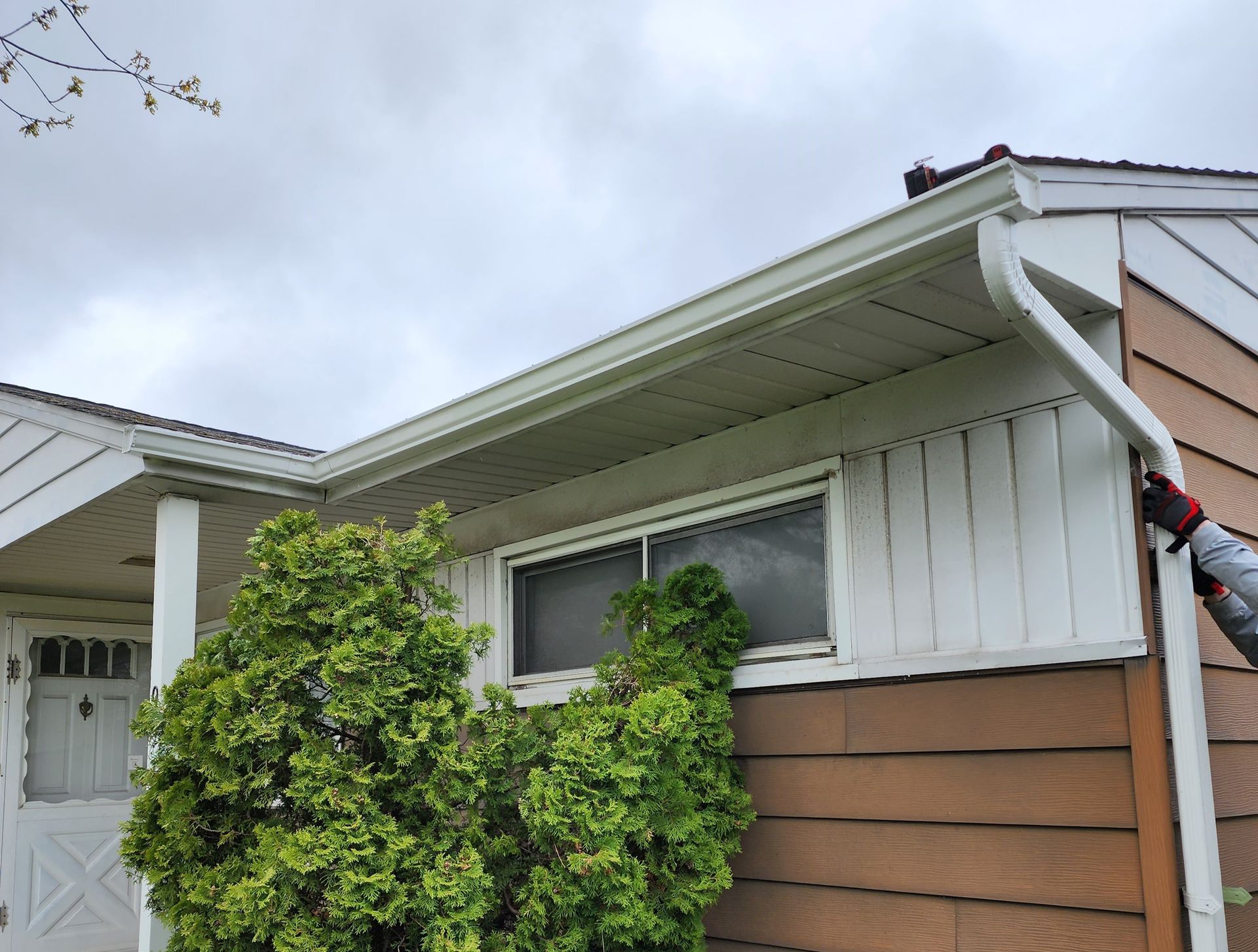 A person’s gloved hand holds a white gutter downspout along the side of a house with tan siding and a large green bush.