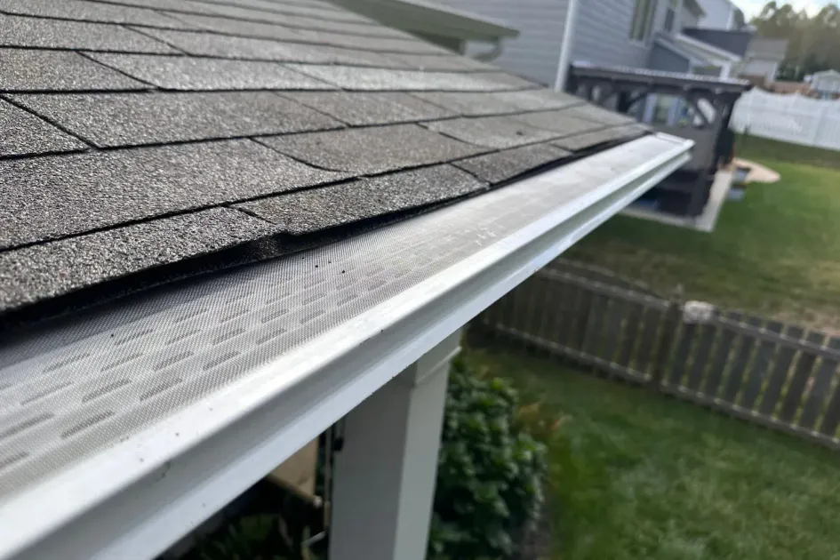A high-angle view of a gray asphalt shingle roof edge with a white gutter covered by a mesh leaf guard.
