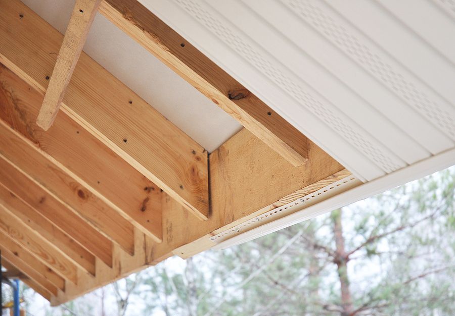 Exposed wooden rafters and joists under a house eave with white vinyl soffit paneling partially installed.