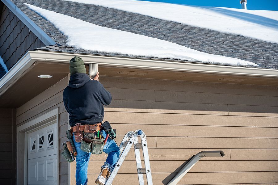 A worker on a ladder installs or repairs a gutter on the side of a tan house with a snow-covered roof.