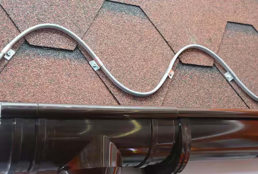 A serpentine heat cable installed in clips along a brown shingled roof edge above a gutter.