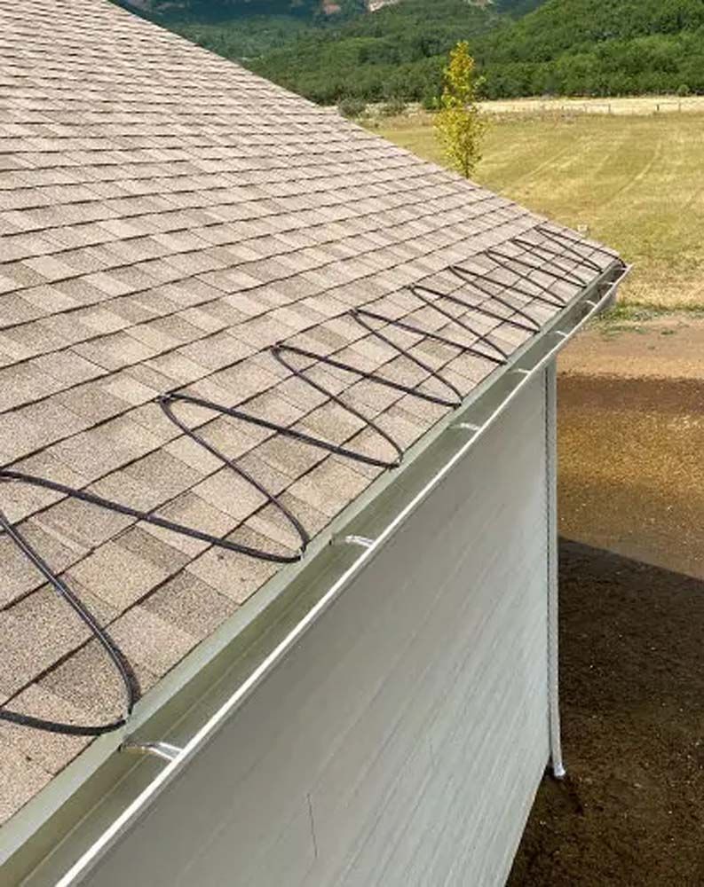 A black zig-zagging heat cable installed on the shingles above a metal rain gutter on a sloped roof.