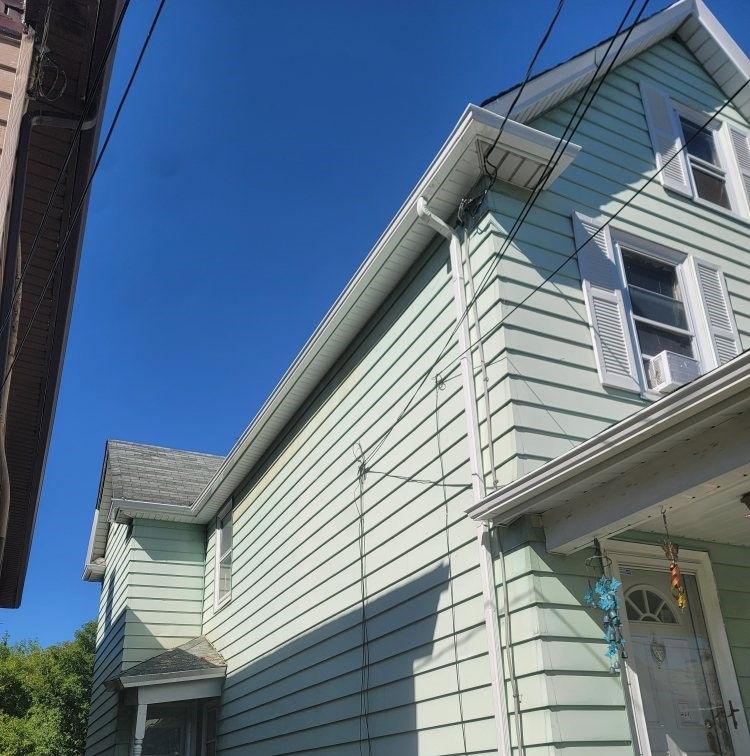 A low-angle view of a light green, two-story wooden house under a bright blue sky on a sunny day.