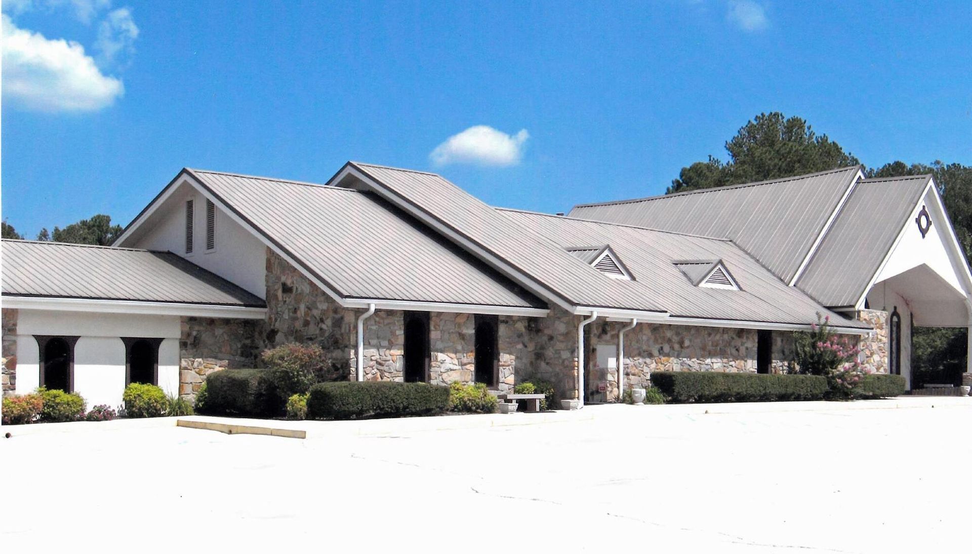 Single-story suburban house with gray metal roofs, stone facade, and white driveway under a blue sky