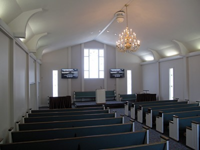 Empty church sanctuary with teal pews, white walls, tall windows, and a chandelier hanging from the ceiling