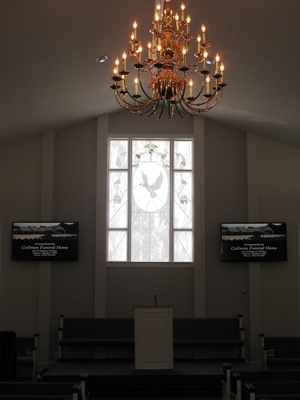 Church sanctuary with chandelier, stained-glass window, stage, and two wall-mounted screens.