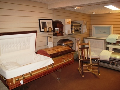 Display of wooden caskets and coffins in a showroom, with a rocking chair and wall shelves.