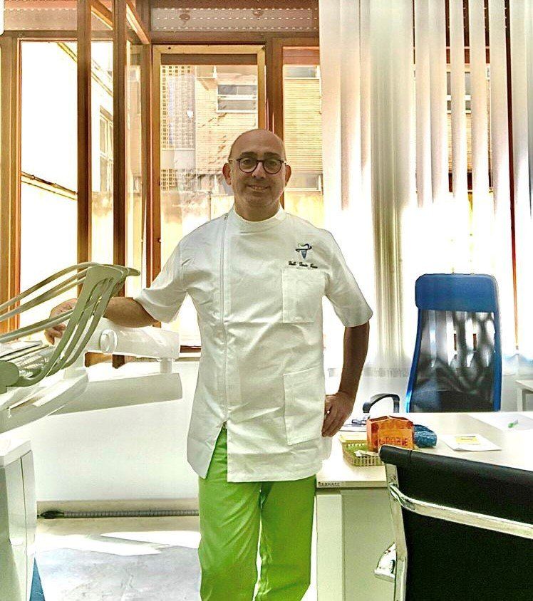 Uomo con giacca da chef bianca e pantaloni verde brillante in piedi in una cucina o sala da pranzo illuminata dal sole.