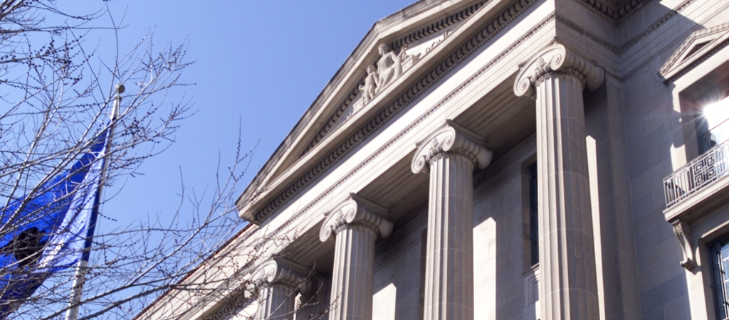 Building with classical columns, a flag, and bare tree branches against a blue sky.