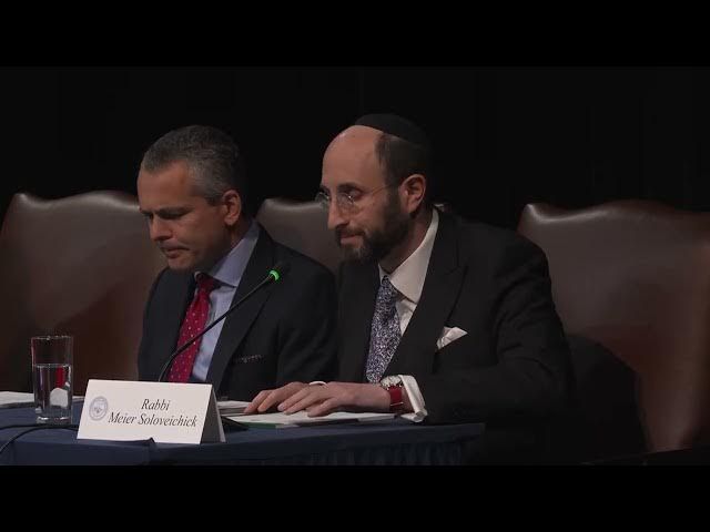 Two men at a table, one with a kippah. Both wear suits, papers, and microphones visible.
