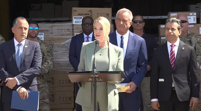 A woman speaks at a podium, surrounded by people in suits and boxes in a warehouse setting.