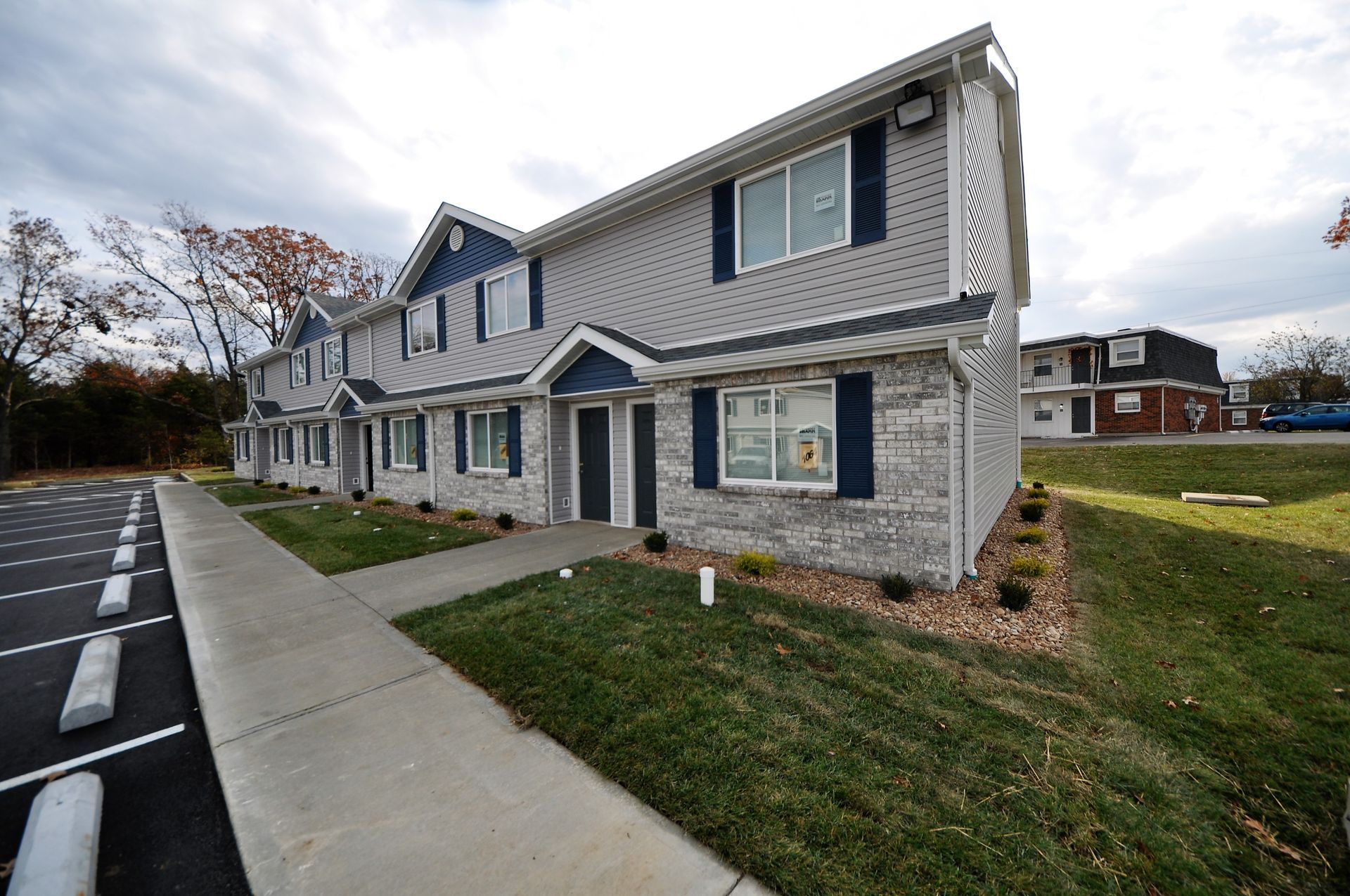 Row of beige townhouses with blue shutters beside a sidewalk and parking lot on a cloudy day