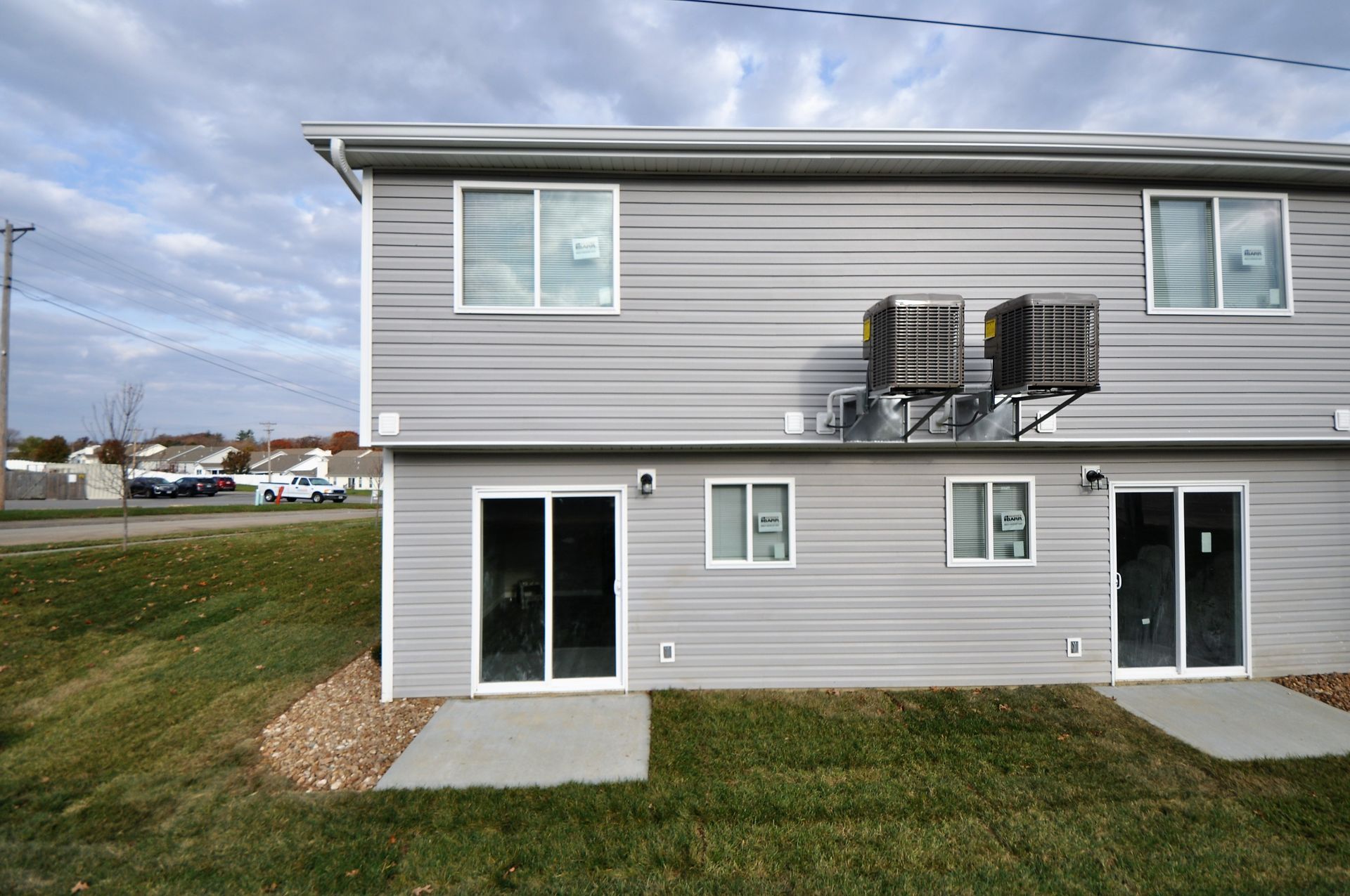 Back of a gray duplex with two patio doors, windows, and wall-mounted air conditioners on a grassy lot