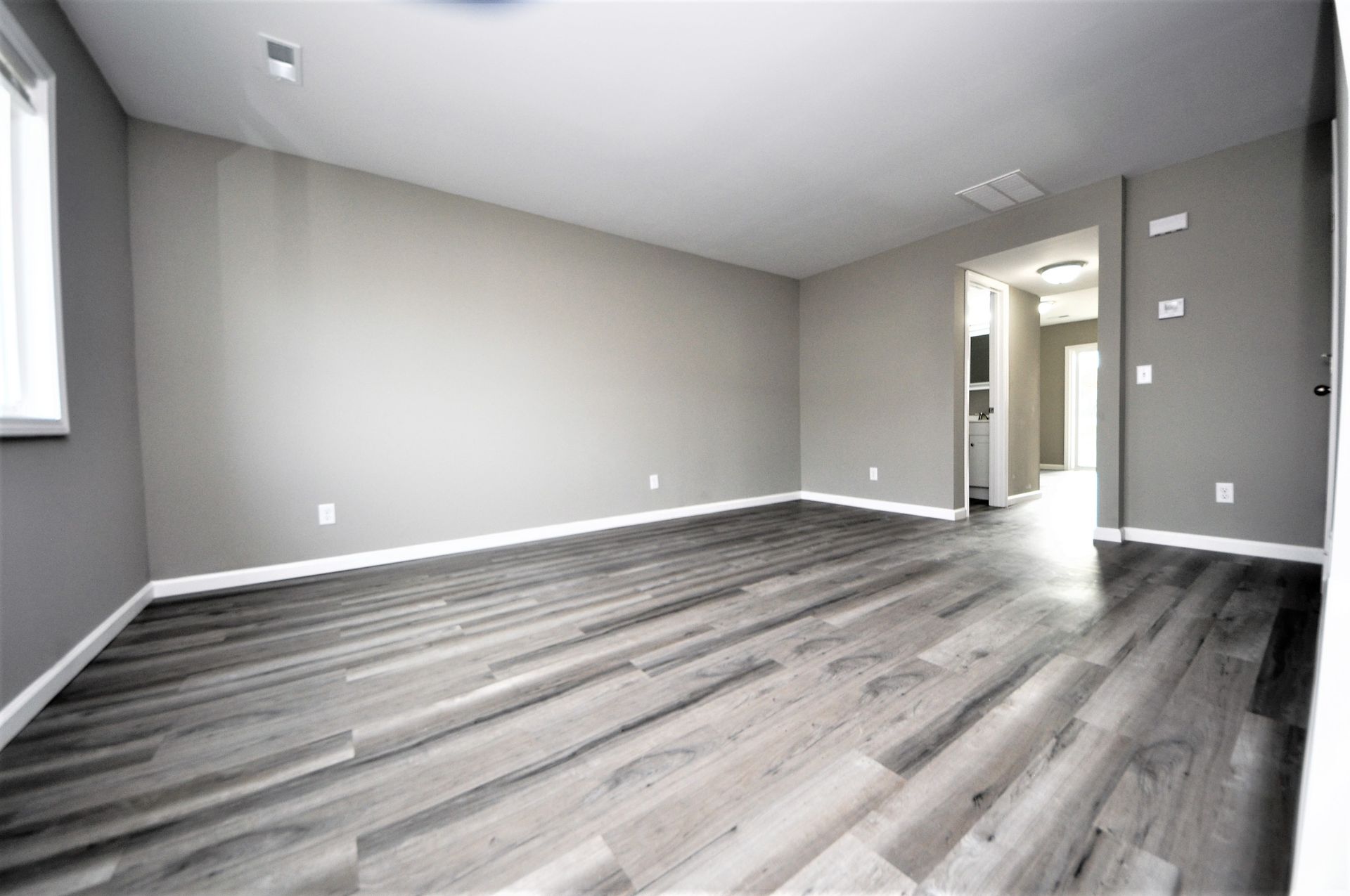 Empty modern room with gray walls, white trim, and gray wood-look flooring, viewed toward a hallway.