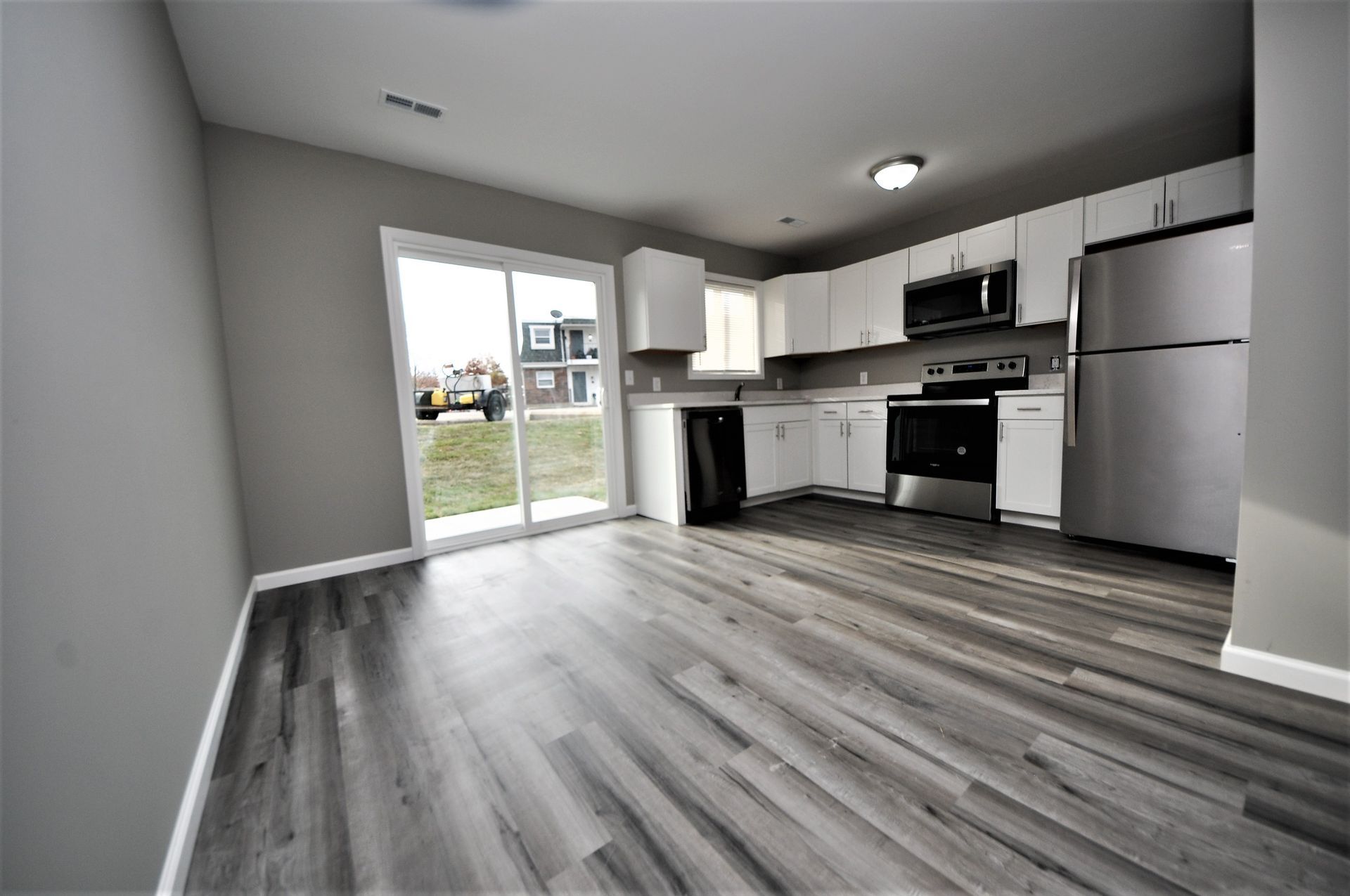 Empty modern kitchen with gray flooring, white cabinets, stainless appliances, and sliding glass door to backyard.