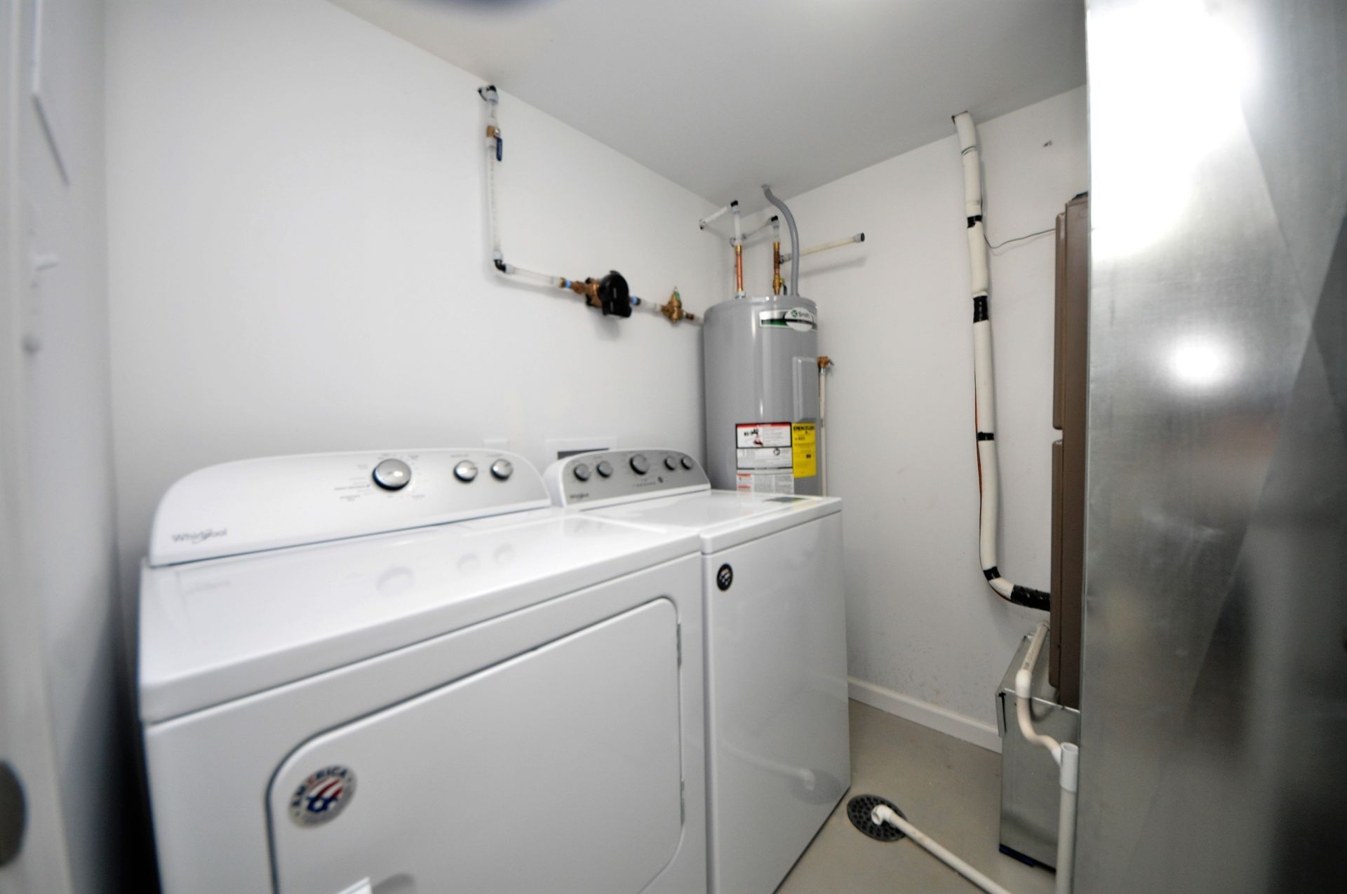 Laundry room with white washer and dryer beside a water heater and utility piping.