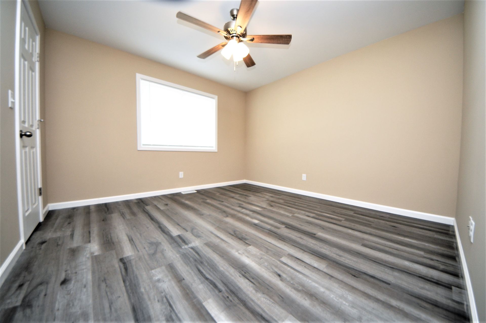 Empty beige bedroom with gray wood-look flooring, a window, and a ceiling fan.