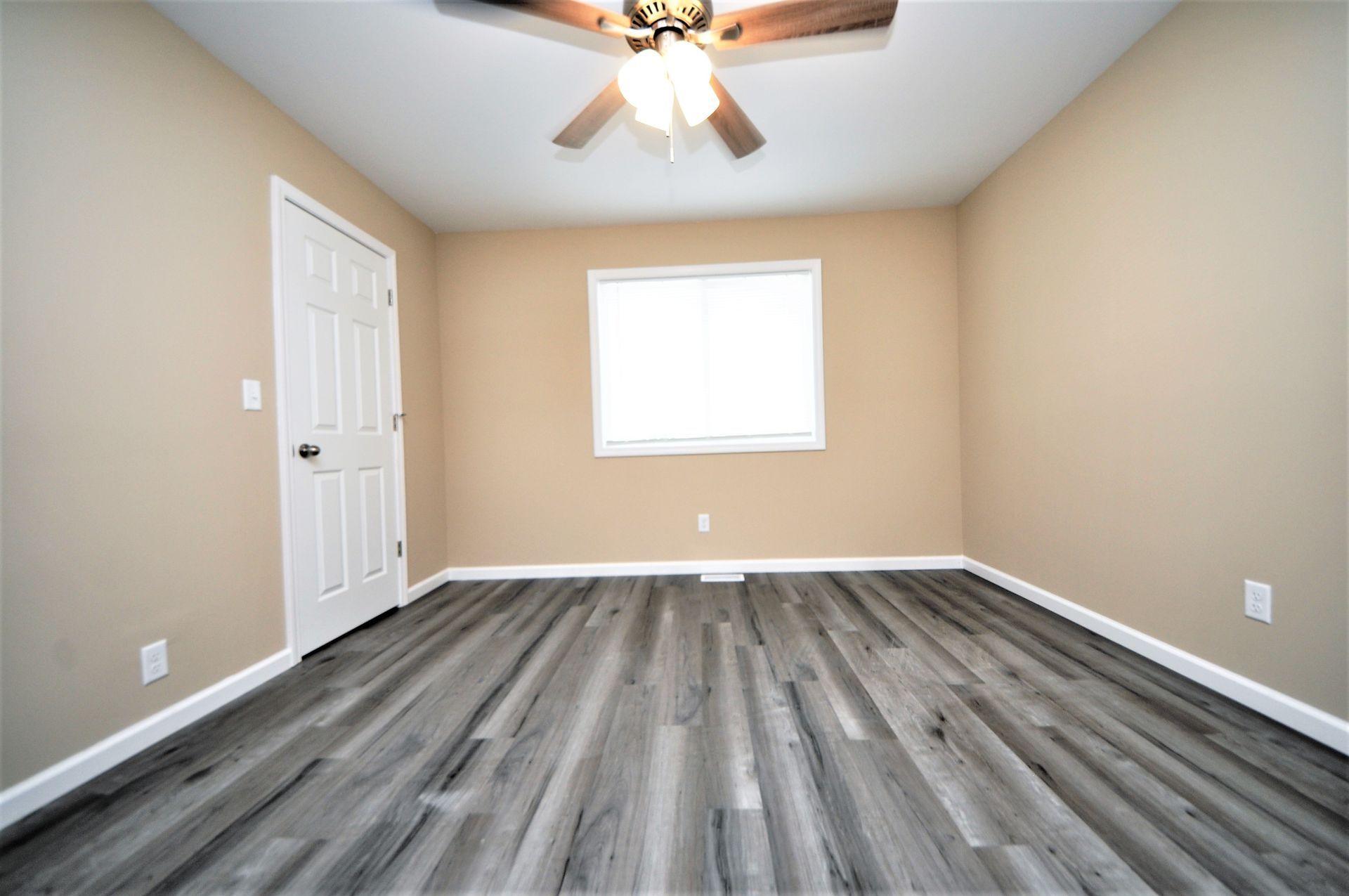 Empty beige bedroom with gray wood-look flooring, ceiling fan, and one window.