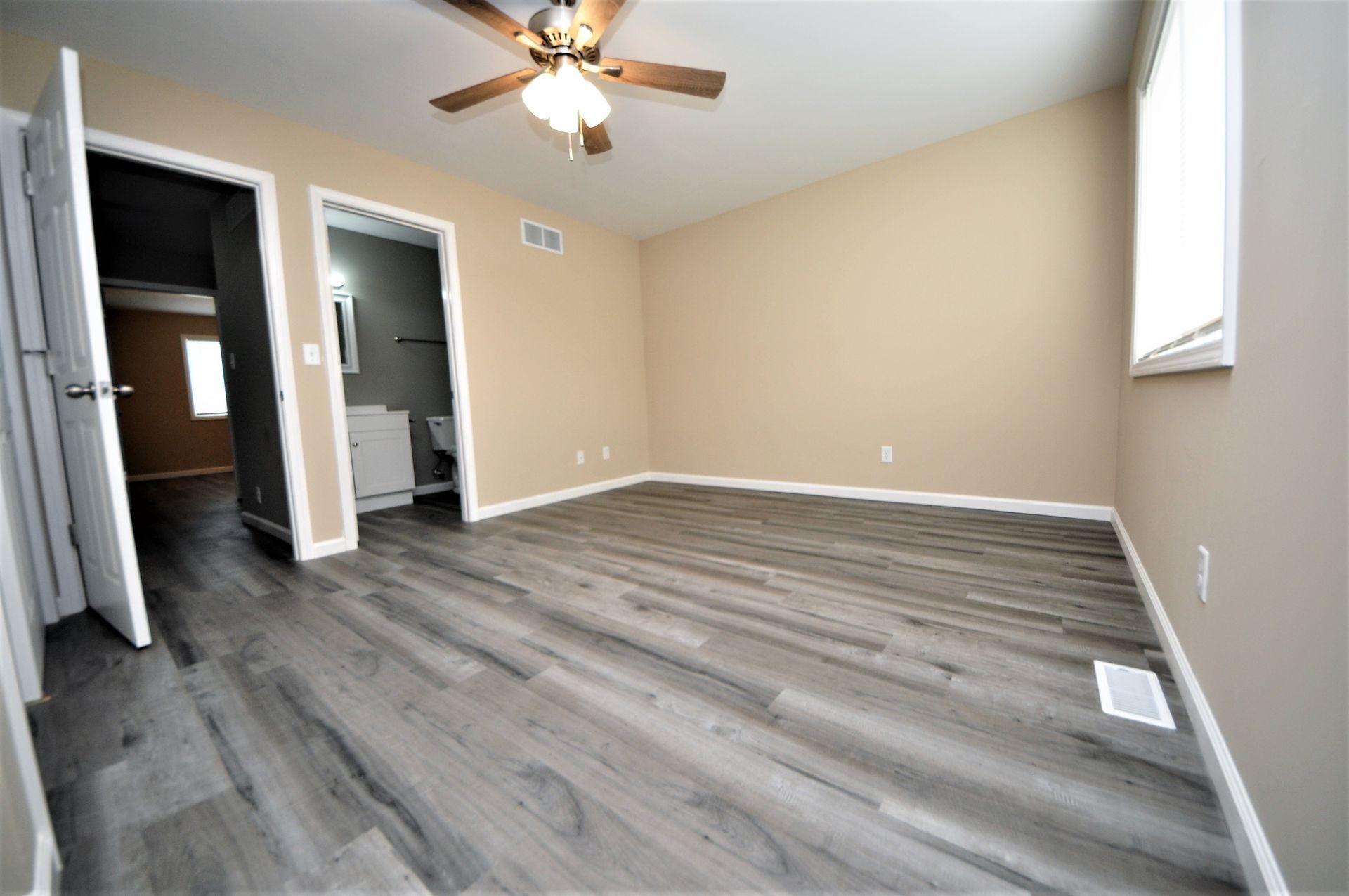 Empty beige bedroom with gray wood flooring, ceiling fan, two doorways, and a window