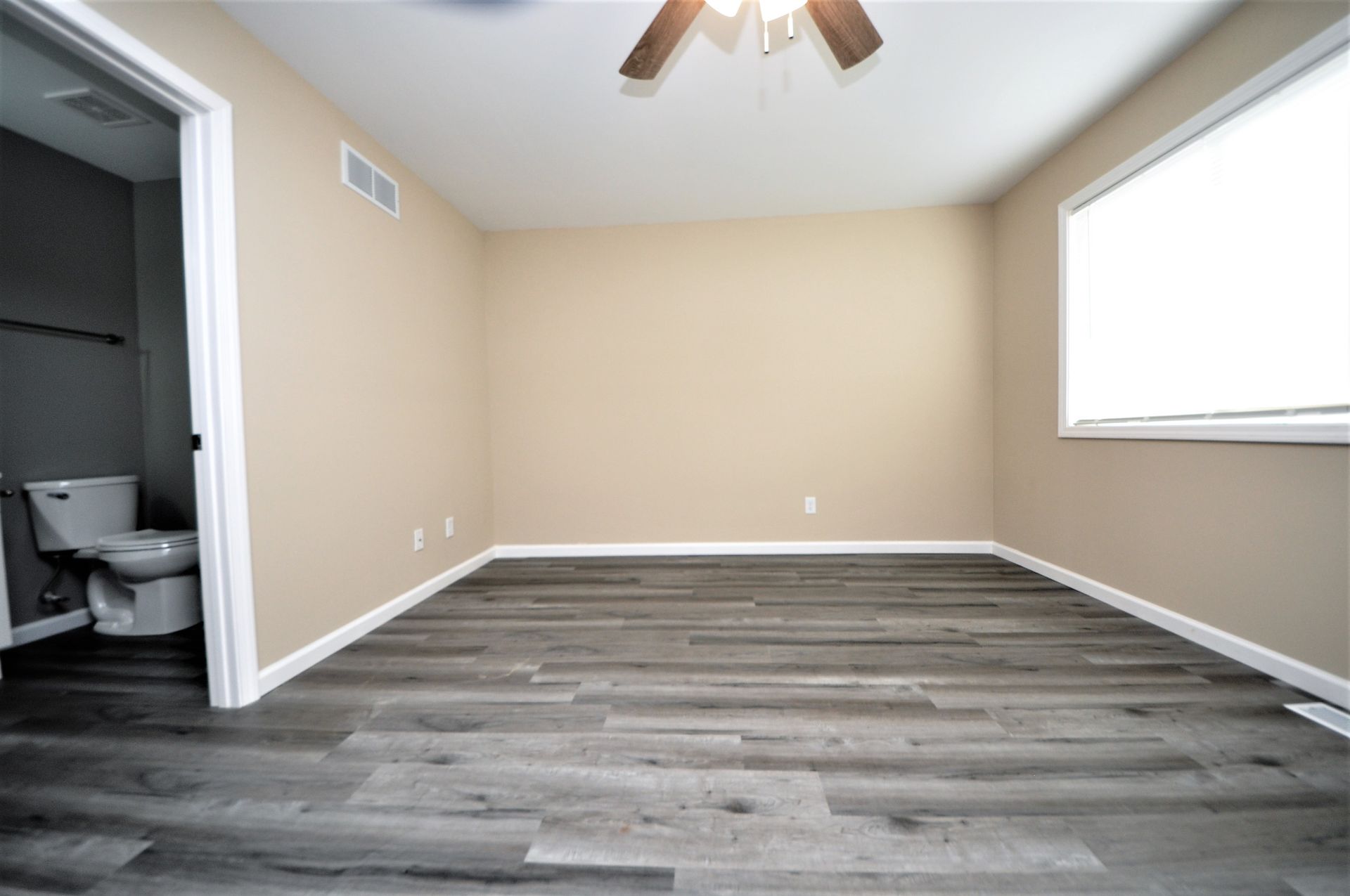 Empty beige bedroom with gray wood-look flooring, a window, ceiling fan, and attached bathroom doorway