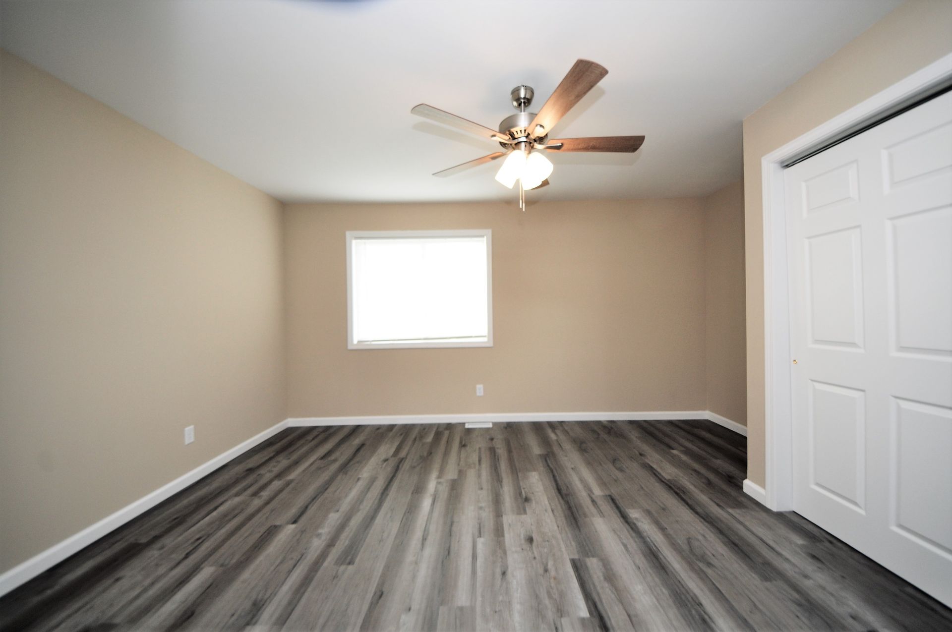 Empty beige bedroom with gray wood flooring, ceiling fan, window, and white closet doors