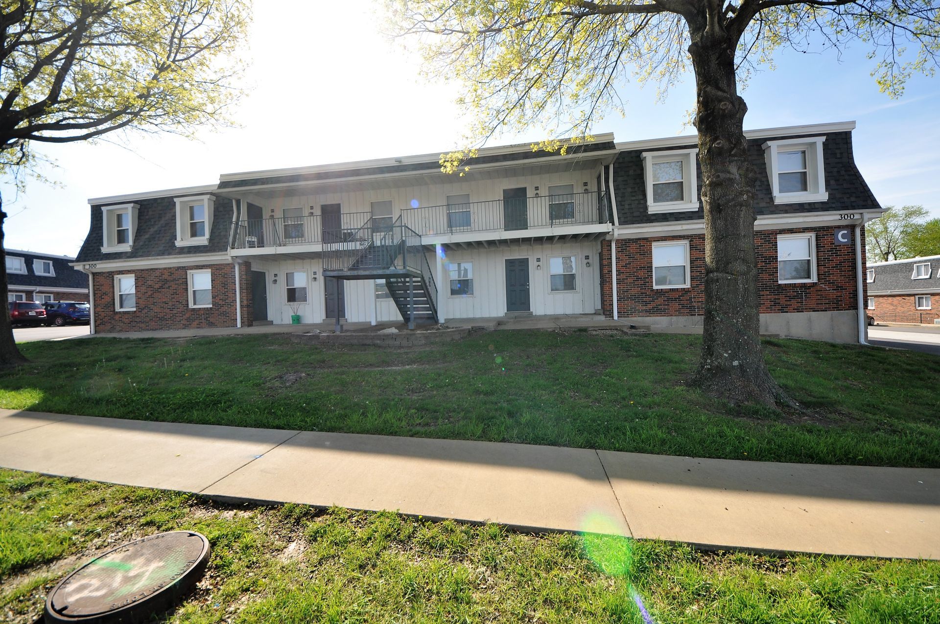 Two-story brick apartment building with green lawn and sidewalk in front, under trees