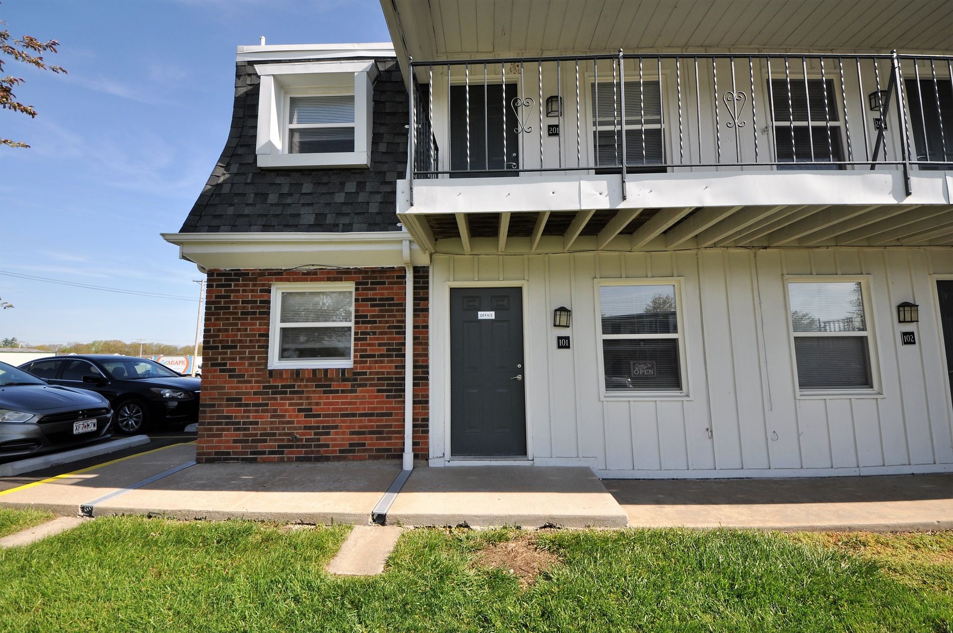 Two-story apartment building with white siding, brick corner, balcony, and parked cars by a grassy lawn
