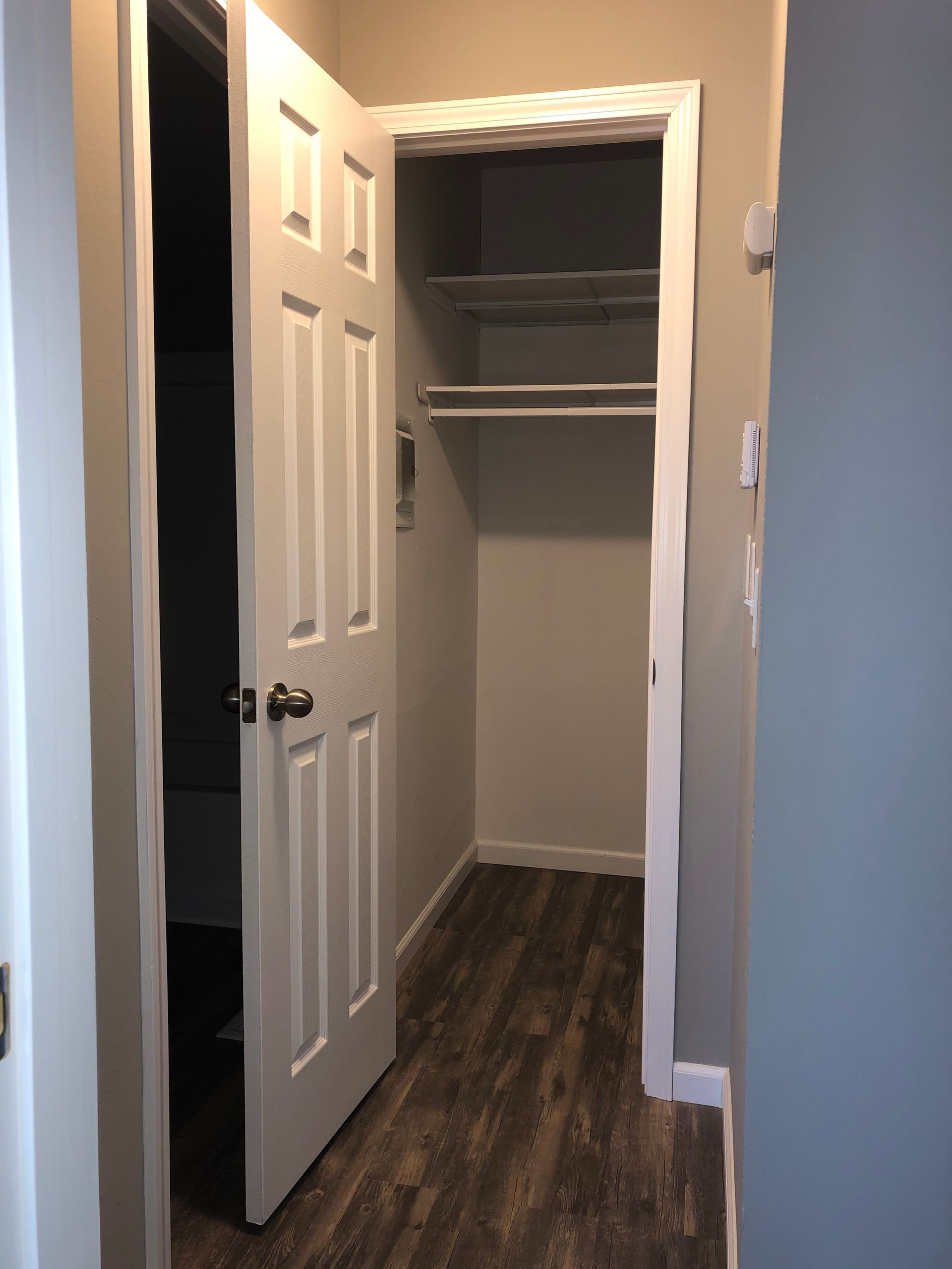 Open closet with white shelves and doors in a hallway, with dark wood flooring.