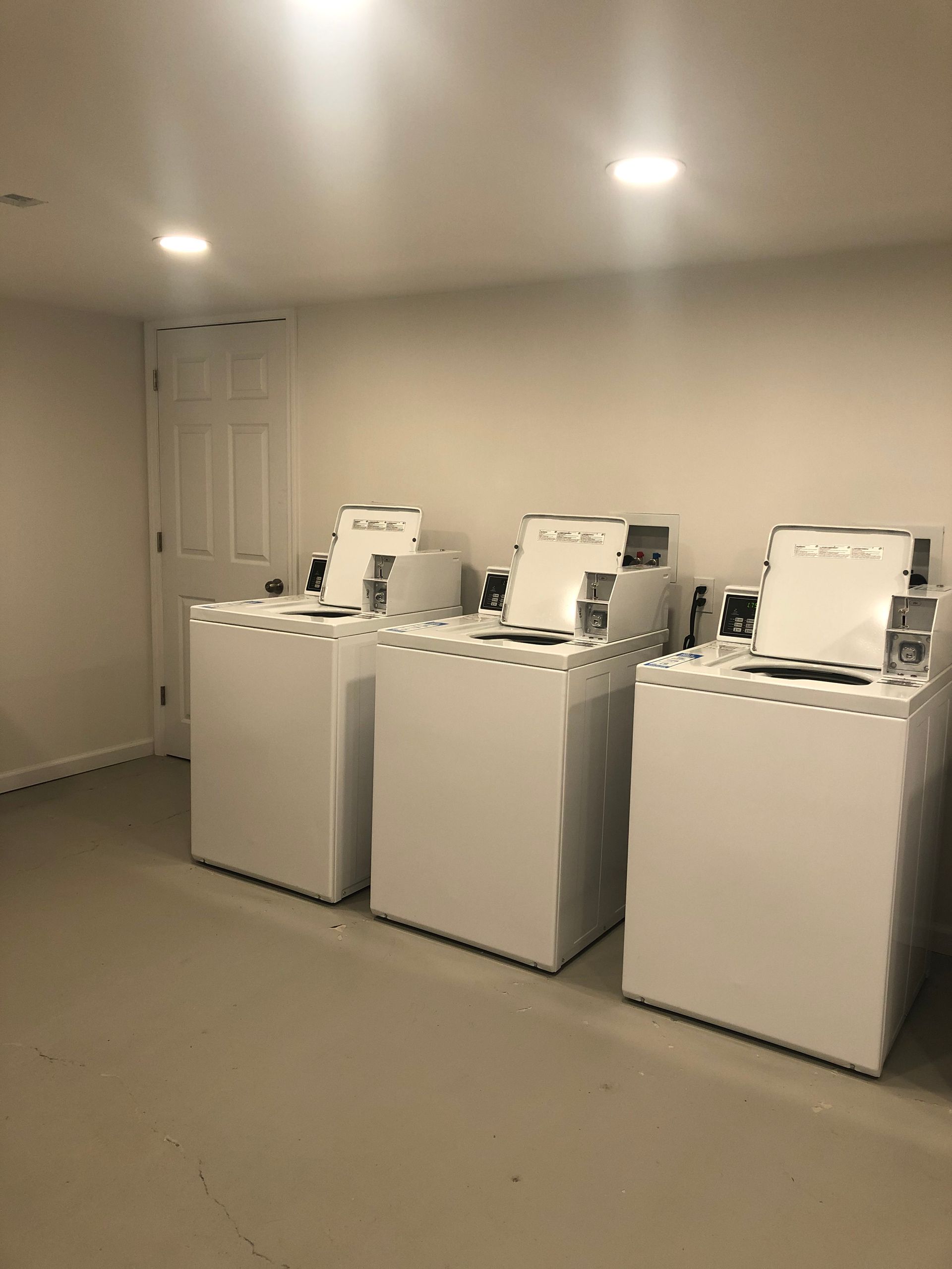 Laundry room with three white top-loading washing machines under recessed ceiling lights.