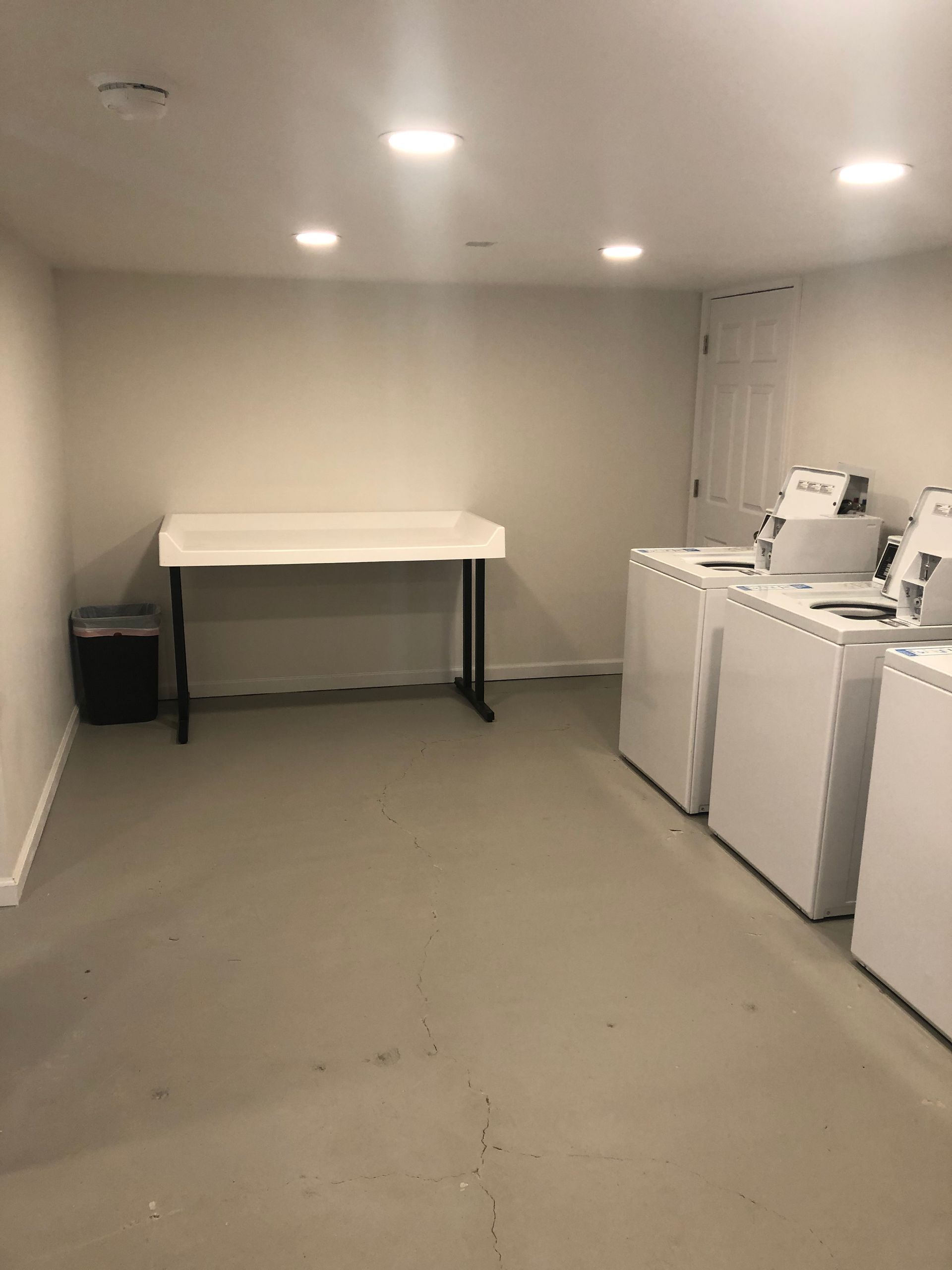 Small utility room with a table, washing machines, and a black bin under recessed ceiling lights.