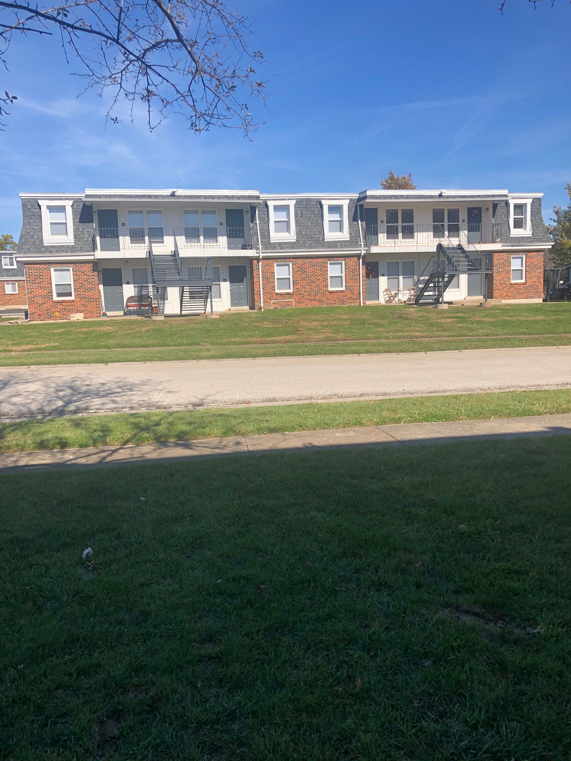 Two-story apartment building with stone and beige siding, dormer windows, and a grassy foreground under a blue sky