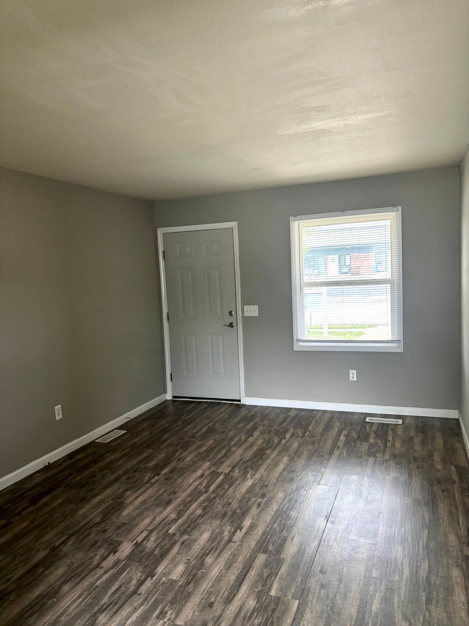 Empty room with gray walls, dark wood floor, white door, and a window letting in daylight.