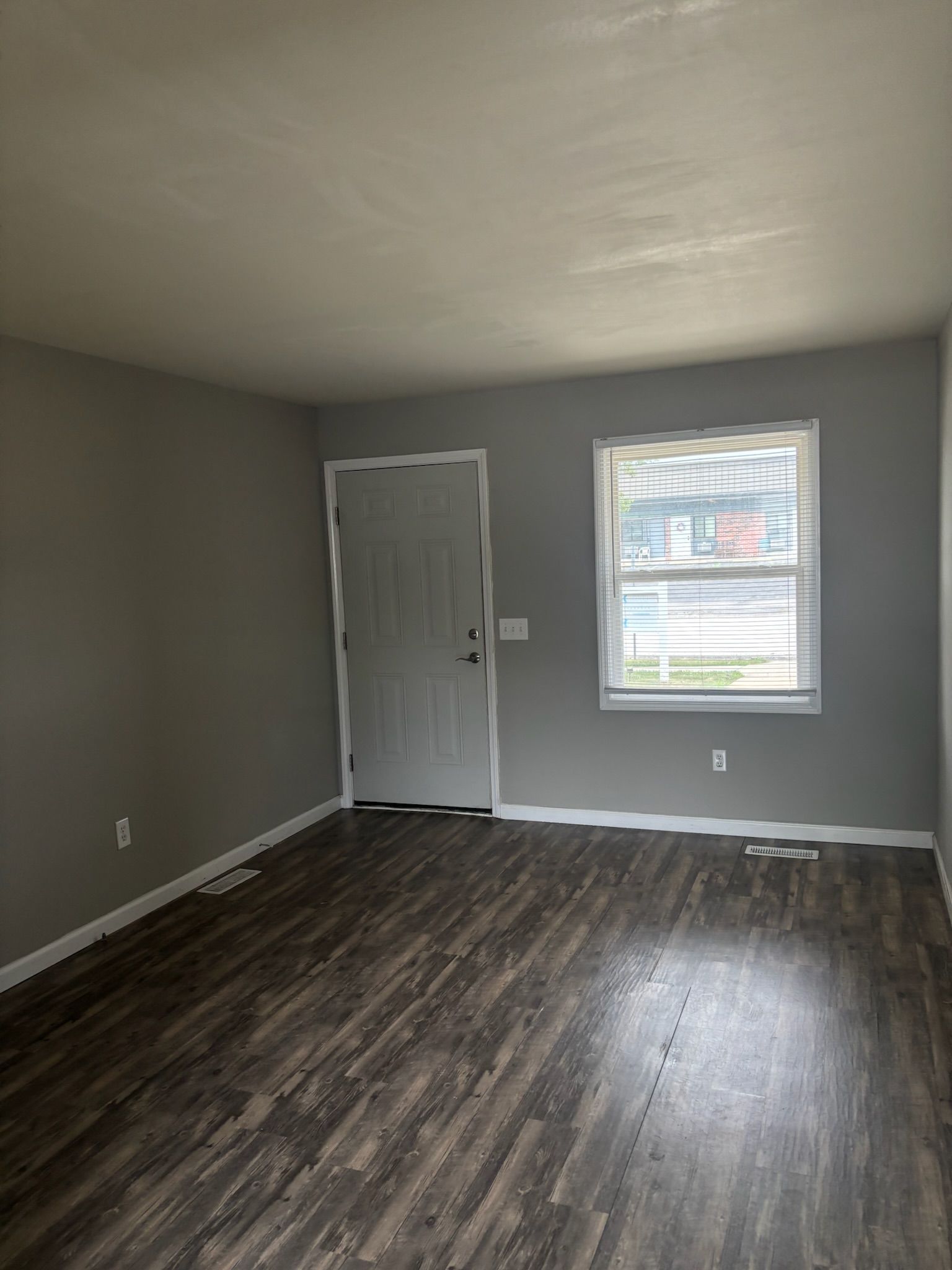 Empty gray room with wood flooring, a white door, and a window letting in daylight