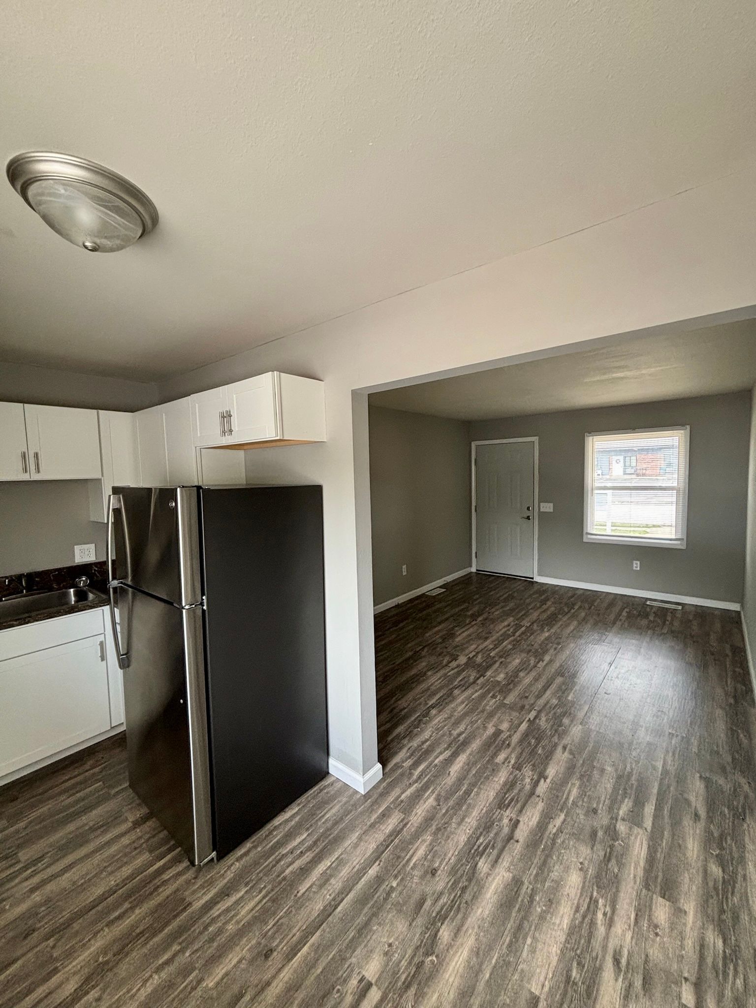 Empty living room and kitchen with dark wood flooring, white walls, and a window in the back.