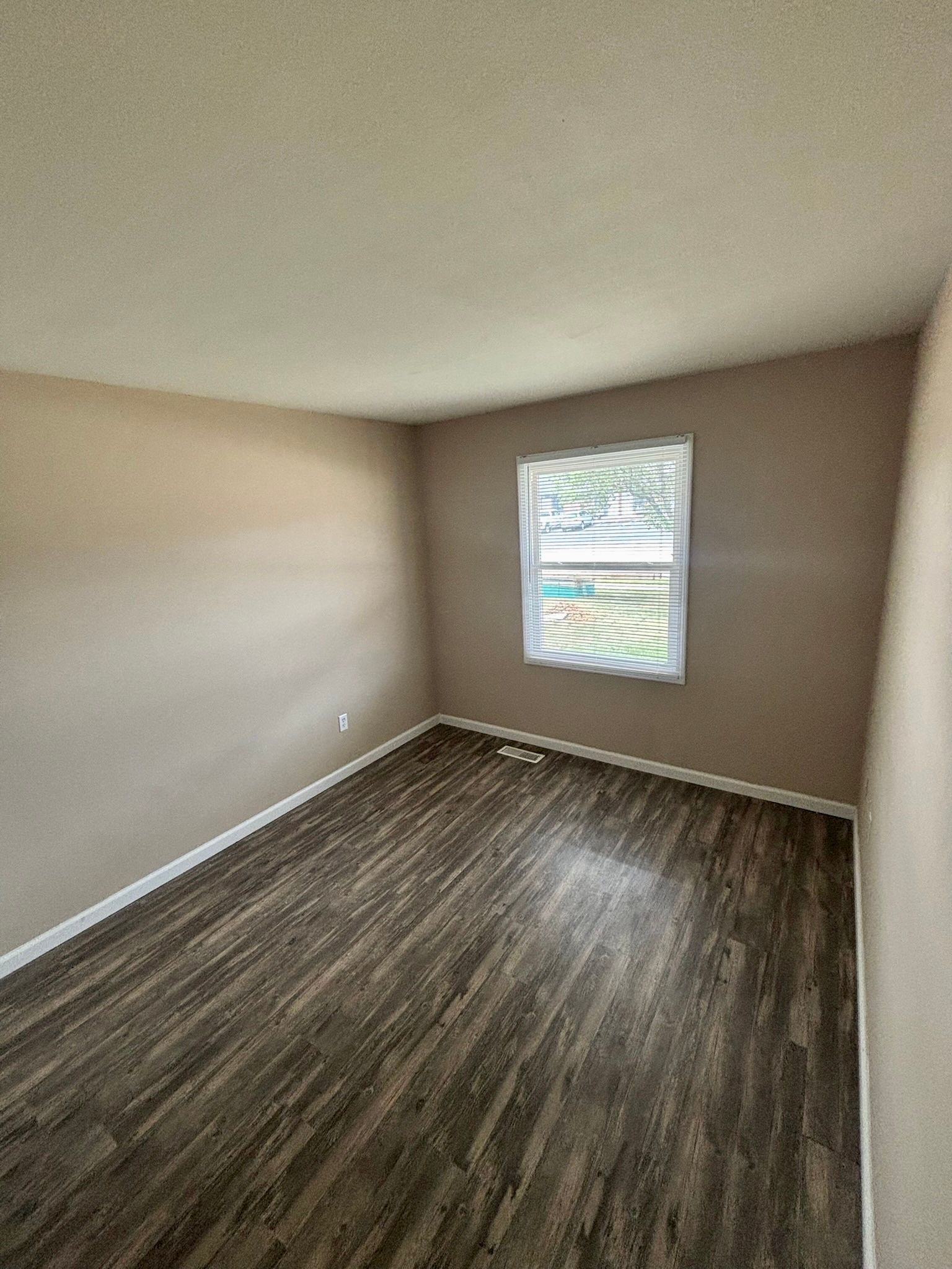 Empty beige bedroom with dark wood flooring and a small window with blinds.