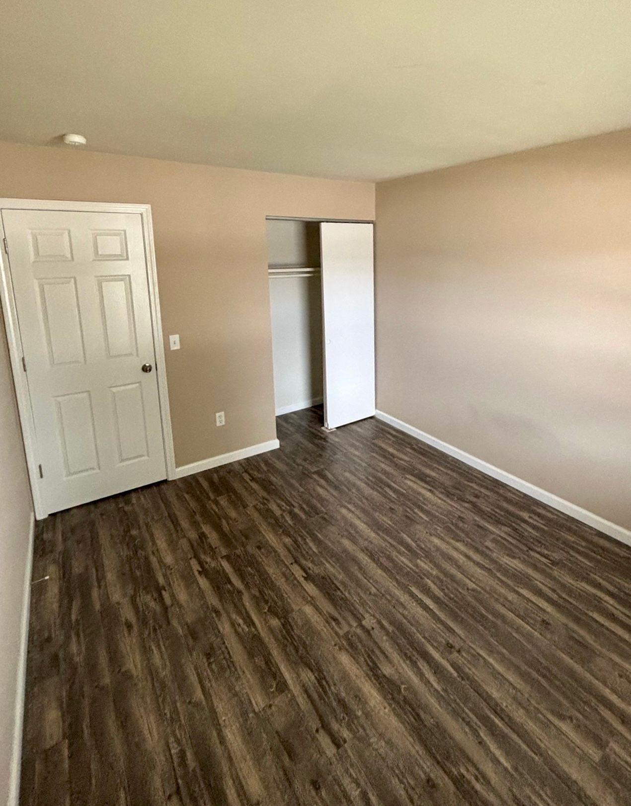Empty beige bedroom with wood-look flooring, white closet doors, and an open doorway.