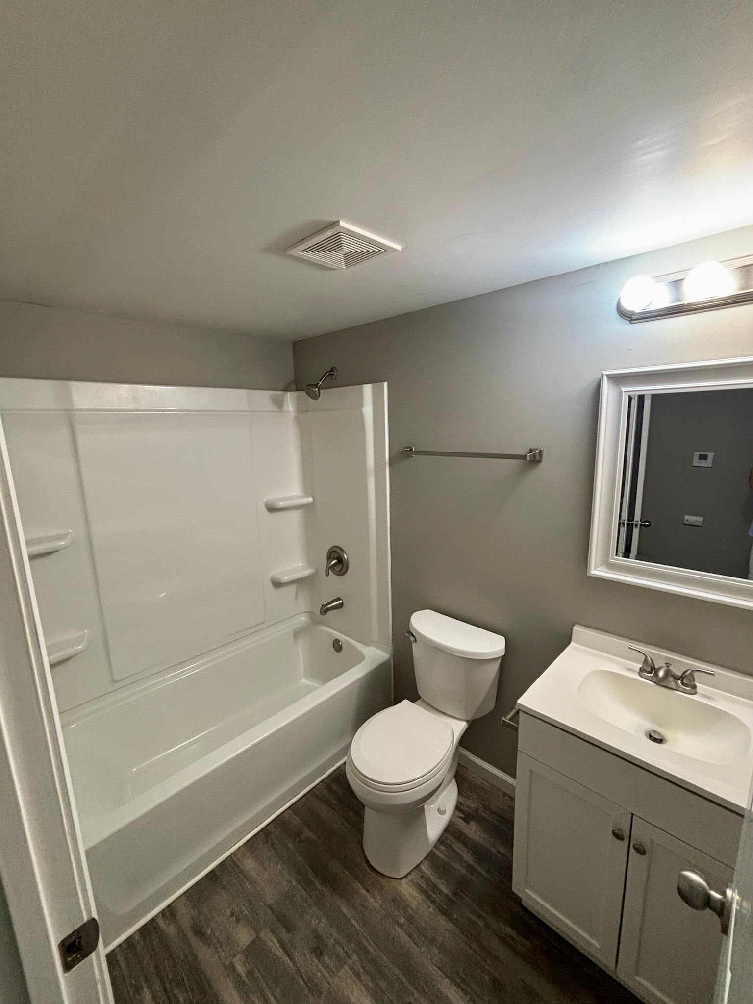 Bathroom with white tub, toilet, and vanity; gray walls and a lighted mirror.
