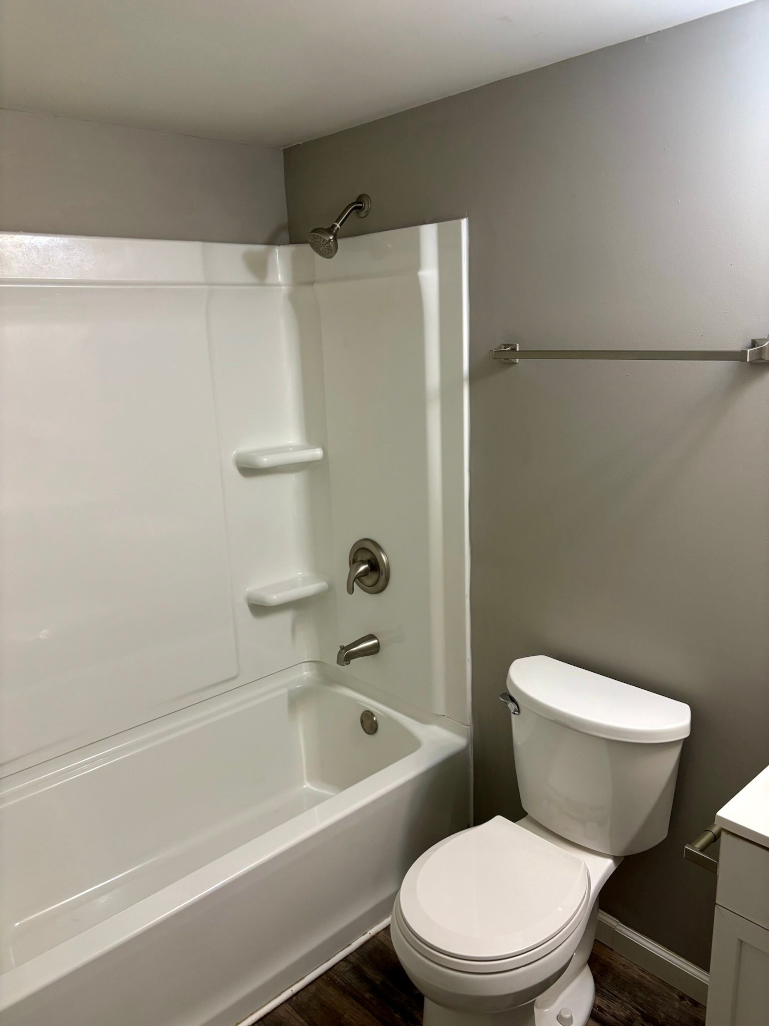 Small bathroom with white tub-shower, toilet, towel bar, and gray walls.