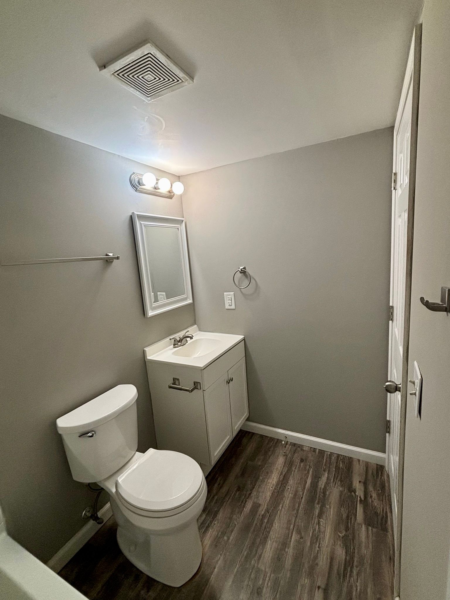 Small beige bathroom with toilet, sink vanity, mirror, towel bar, and dark wood-look floor.
